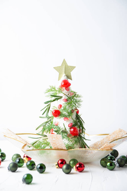 Vista del árbol de queso con eneldo, romero, cerezas y granada, con decoraciones navideñas, sobre mesa blanca con bolas rojas y verdes, fondo blanco, vertical, con espacio para copiar - Foto, Imagen