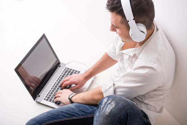 Young man is sitting on the floor with laptop and headphone. Grey background. - Фото, изображение
