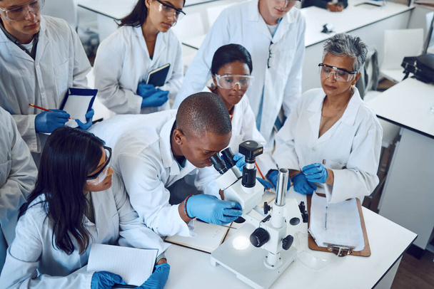Science, students and medical laboratory with microscope and mentor for pharmaceutical research, education and learning in class. Scientific and chemistry men and women group doing analysis of drug. - Photo, Image