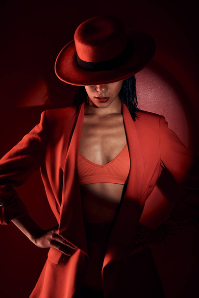 Woman, fashion hat and spotlight for suit, sexy and mystery in studio for beauty, punk aesthetic and cosmetics. Fashion model girl, cyberpunk design or unique with dark cosmetic by red background. - Photo, Image