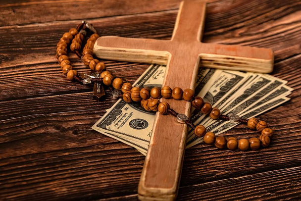 Een katholiek kruis, een rozenkrans met kralen en dollars liggen op een donkerbruine houten tafel. - Foto, afbeelding