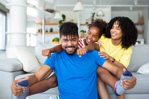 Happy African American dad and mom with excited proud daughter kid, playing flying superhero, reaching arm forward. Cheerful girl playing active game with family at home - Photo, Image