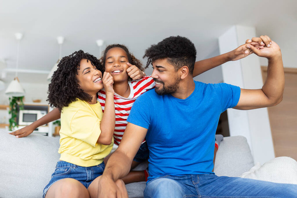 Glückliches Familienporträt. Fröhliche Mutter, Vater und ihre süße Tochter posieren zu Hause im Wohnzimmer, kleines Mädchen sitzt auf Papas Schultern, freier Raum - Foto, Bild