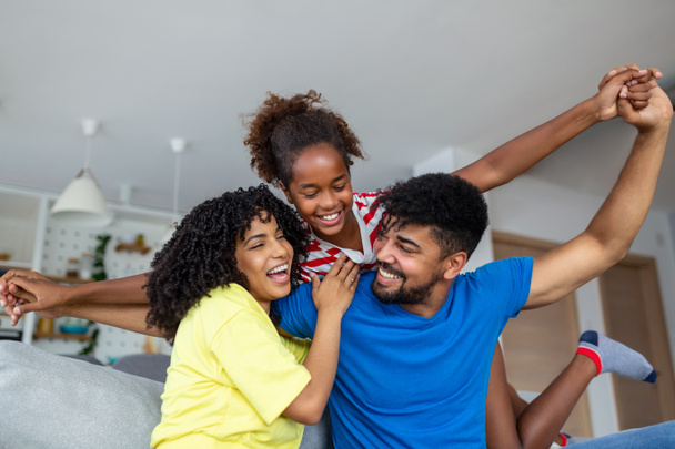 Happy African American dad and mom piggybacking excited proud daughter kid, playing flying superhero, reaching arm forward. Cheerful girl riding fathers back playing active game with family at home - Photo, Image