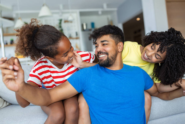 Attraktive glückliche Familie umarmt attraktive Mutter schöner Vater und entzückende kleine Tochter sitzen zusammen auf der Couch im Wohnzimmer zu Hause lächelnd in die Kamera - Foto, Bild