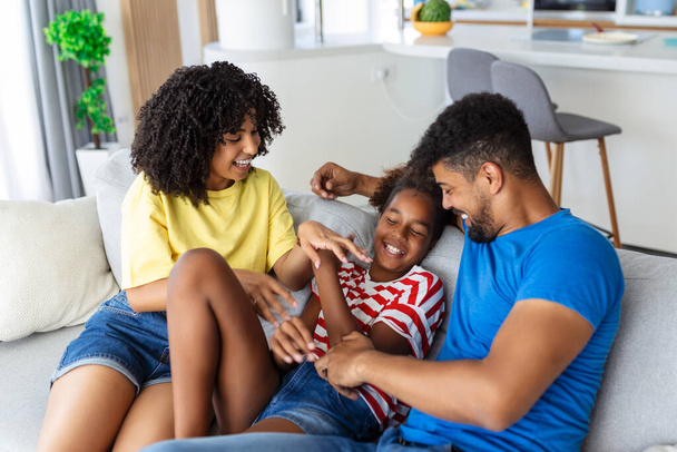Feliz familia multiétnica sentados en el sofá riendo juntos. Padres alegres jugando con su hija en casa. Padre hace cosquillas a su niña mientras la madre sonríe. - Foto, imagen