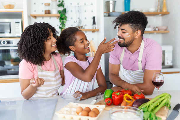 Menina bonito e seus belos pais estão sorrindo enquanto cozinha na cozinha em casa. Família afro-americana feliz preparando comida saudável juntos na cozinha - Foto, Imagem