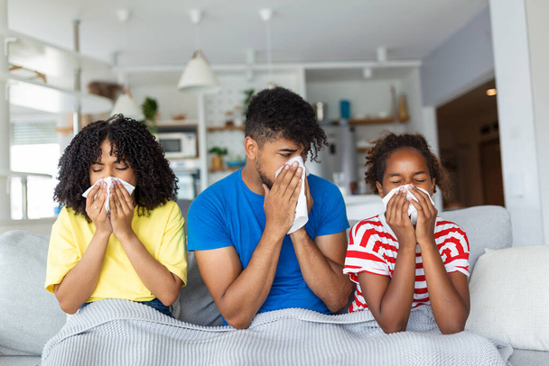Concepto Gripe y Enfermedad. Retrato de una joven familia afroamericana enferma sonándose las narices con servilletas juntas mientras están sentados en el sofá cubierto con manta. Cuatro personas enfermas con frío en casa - Foto, Imagen