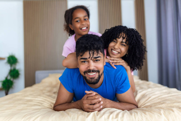 Portrait de belle jeune mère, père et leur fille regardant la caméra et souriant tout couché sur le lit appuyé sur l'autre
 - Photo, image