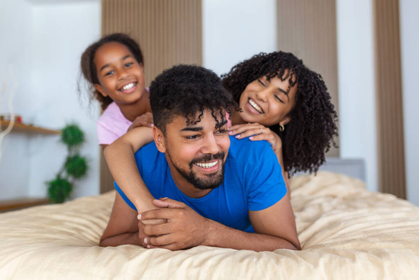 Uśmiechnięta afrykańska rodzina trojga leżąca na łóżku, jeden na górze, jeden w środku i jeden na dole.. - Zdjęcie, obraz