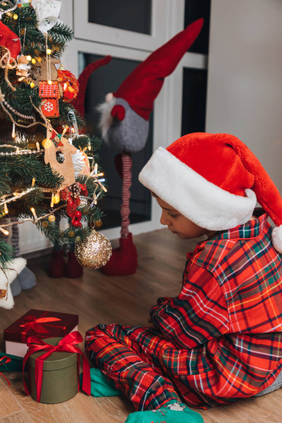 Un ragazzo in pigiama rosso e cappello di Babbo Natale si siede vicino a un albero di Natale con regali a casa. Natale e famiglia. Vista frontale - Foto, immagini