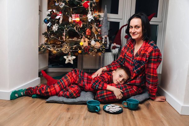 Maman et son fils en pyjama rouge s'assoient près du sapin de Noël avec du thé et des biscuits au chocolat. Vue de face - Photo, image