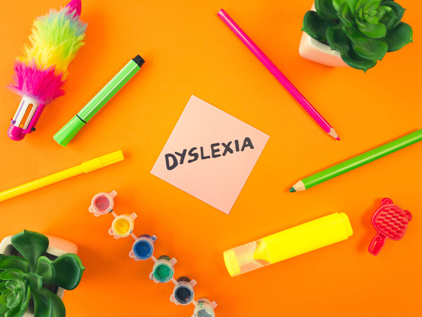 Слово "дислексия" написано от руки на липкой ноте с детскими принадлежностями на оранжевом фоне. Сложность обучения, концепция образования - Фото, изображение