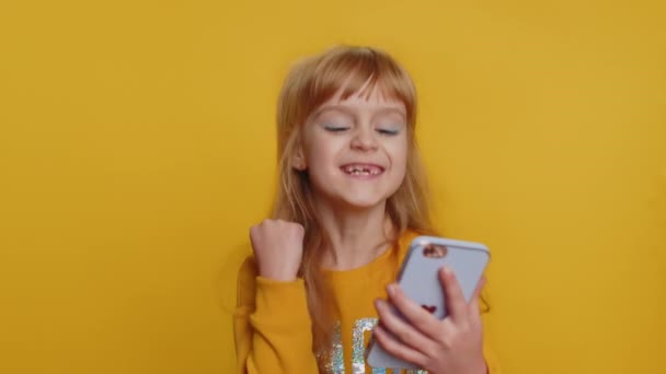 Young preteen child girl kid use mobile smartphone typing browsing say Wow yes found out great big win good news celebrate doing winner gesture. Little toddler children on studio yellow background - Filmati, video