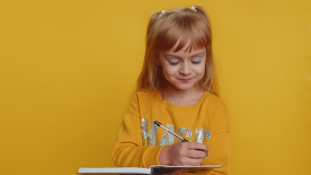 Thoughtful young preteen child girl kid making notes, writing down thoughts with pen into notepad notebook diary, to do list, good idea. Little toddler children pupil isolated on yellow background - Footage, Video