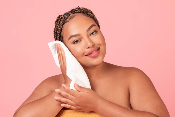 Hygienekonzept. Schöne schwarze Plus-Size-Frau trocknet sauberes Gesicht mit Handtuch, steht über rosa Studiohintergrund. Schönheitsroutine am Morgen - Foto, Bild