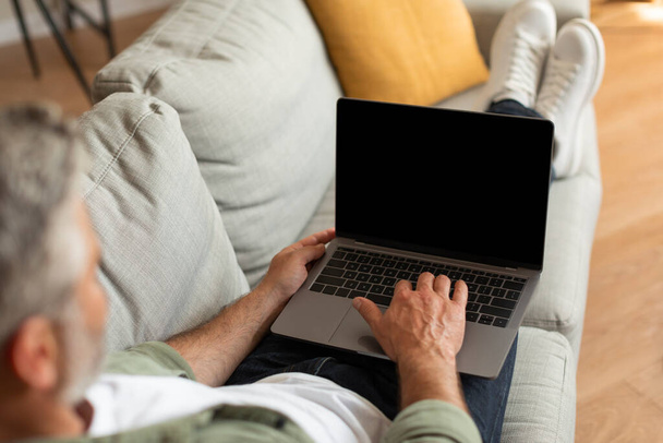 Ostříhaný dospělý běloch, který píše na počítači s prázdnou obrazovkou, odpočívá, leží na pohovce a sleduje video v obývacím pokoji, zblízka. Technologie pro práci a setkání, reklama a nabídka, relaxace doma - Fotografie, Obrázek