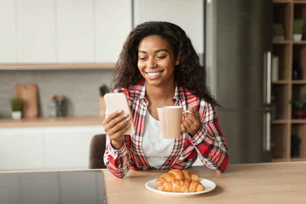 Glimlachende duizendjarige zwarte dame met kopje koffie drinken, Bekijk video op de smartphone, heeft videogesprek, genieten van het ontbijt in de keuken interieur. Goedemorgen, ontmoeting op afstand, communicatie met technologie thuis - Foto, afbeelding