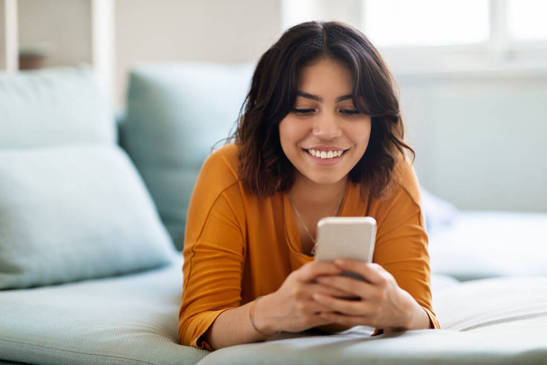 Mobilfunk. Fröhliche arabische Botschaften auf dem Smartphone, während man zu Hause auf der Couch liegt, lächelnde junge Frau aus dem Nahen Osten, die mit Freunden SMS schreibt oder in sozialen Netzwerken surft - Foto, Bild