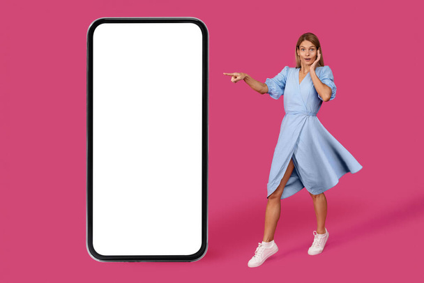 Shocked adult caucasian lady blonde in dress point finger at big smartphone with blank screen, isolated on pink background, studio. Online surprise, great news, sale, online app for blog, ad and offer - Photo, Image