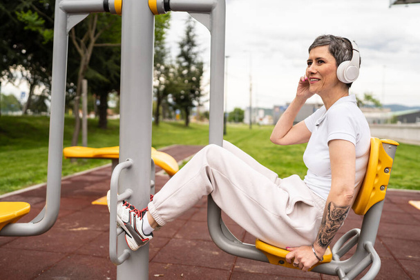 Una mujer madura o mayor caucásica entrenamiento femenino en la prensa de piernas máquina de fitness urbana en el parque en el gimnasio al aire libre Deporte concepto de estilo de vida saludable personas reales copiar espacio - Foto, imagen