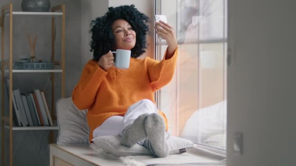Афроамериканская женщина позирует для фото с чашкой горячего чая в гостиной внутренней замедленной съемки. Счастливая женщина пьет кофе перед камерой телефона у окна. Красавица снимает видео на смартфон - Кадры, видео