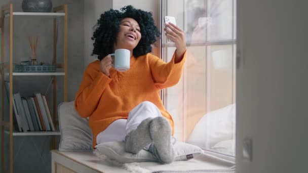 Mulher feliz bebendo café na frente da câmera do telefone na janela. Menina bonita fazendo vídeo no smartphone. Mulher afro-americana posando para foto com xícara de chá quente na sala de estar interior câmera lenta - Filmagem, Vídeo