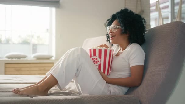 Heureuse Afro-Américaine se relaxant seule sur coach regarder la télévision à la maison. Modèle ethnique joyeux avec des cheveux bouclés foncés riant à haute voix sur le canapé à la maison en regardant un film avec panier de maïs soufflé 4K - Séquence, vidéo