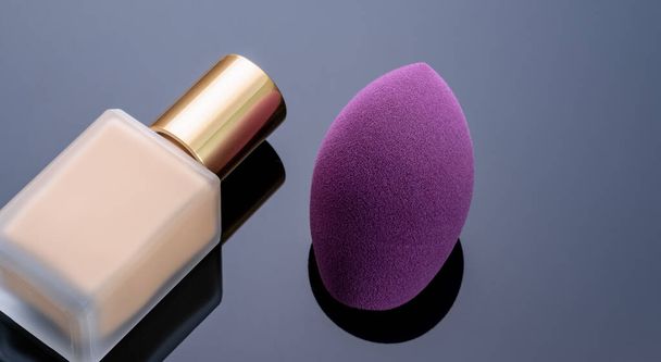 Purple makeup sponge on dark background. Soft makeup cosmetic sponge. Flat-ended makeup sponge with blur makeup foundation bottle. Liquid beige make-up foundation and beauty blender. Cosmetic product. - Photo, Image