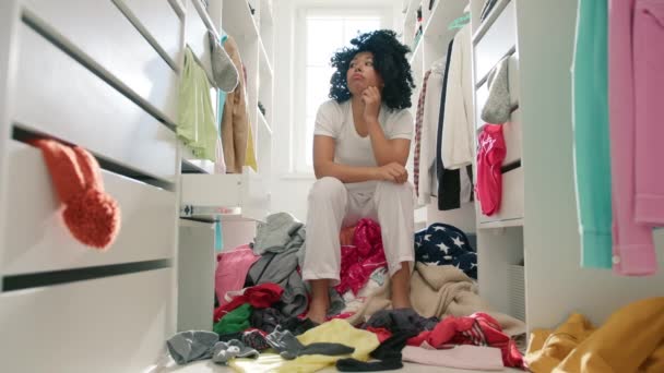 Een donkere vrouw die in de kast loopt. Triest Afro-Amerikaans model omringd door ontvouwde kleren die moe zijn van het opruimen van garderobe. Grote schoonmaakdag. Andere kleding, chaos thuis. - Video