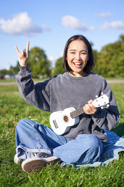 Feliz chica asiática jugando ukelele en parque, mostrando rock n roll, cuernos de metal pesado signo y sonriendo, divertirse al aire libre. - Foto, imagen