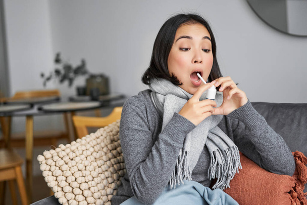 Mujer enferma usando aerosol de dolor de garganta, tratando su resfriado o gripe, quedarse en casa con licencia por enfermedad, sentado en el sofá en la sala de estar. - Foto, imagen