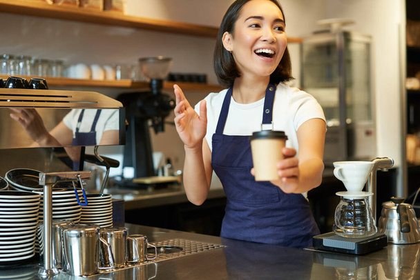 Portret van lachende Aziatische meisje barista, het geven van orde in cafe, uitnodigen van gasten op te halen afhaalbon in de buurt van toonbank, het houden van afhaalkopje koffie. - Foto, afbeelding