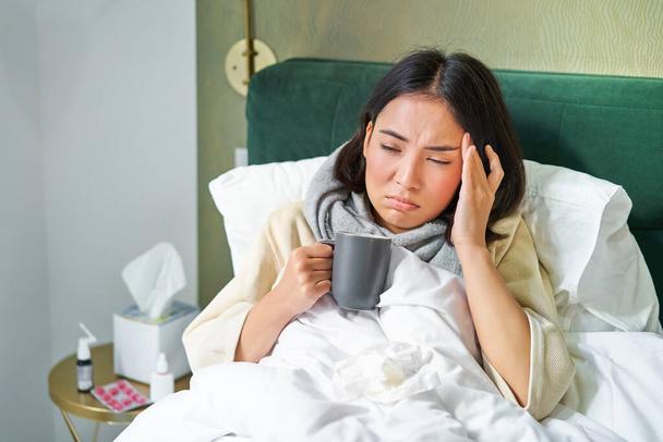 Retrato de una mujer asiática con dolor de cabeza, cogiendo col, permaneciendo en licencia por enfermedad en casa, acostada en la cama, bebiendo té caliente, teniendo gripe. - Foto, Imagen