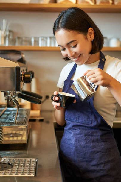 Close up of cute asian barista girl making cappuccino, doing latte art in cup with steamed milk, standing in coffee shop behind counter. - Photo, Image