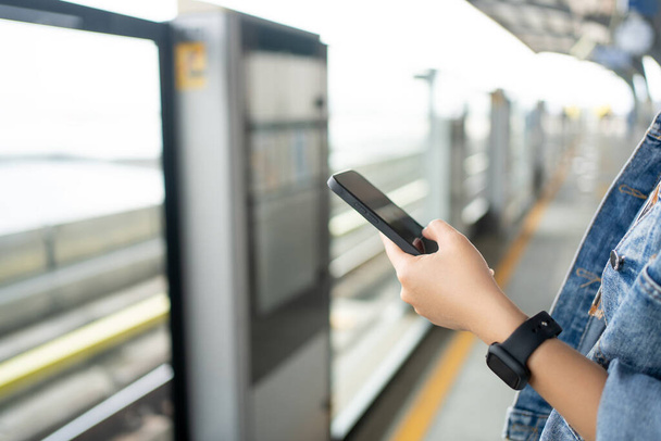 Κοντινό πλάνο του χεριού με τη χρήση smartphone στην πλατφόρμα αναμονής για τη μεταφορά του τρένου ουρανό. - Φωτογραφία, εικόνα