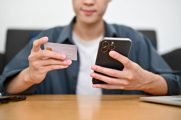 immagine ritagliata, Un bel giovane asiatico siede al tavolo con una carta di credito e uno smartphone. mobile banking, pagamento online, shopping online - Foto, immagini