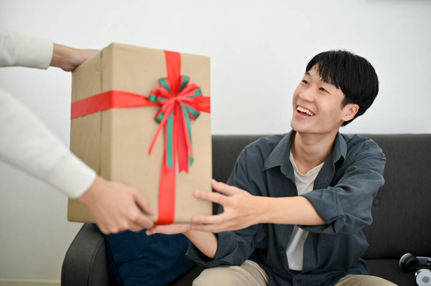 Gelukkige en opgewonden jonge Aziatische man ontvangt een verrassing geschenkdoos van zijn vriend. verjaardagsfeestje, jubileum, bijzonder moment. - Foto, afbeelding