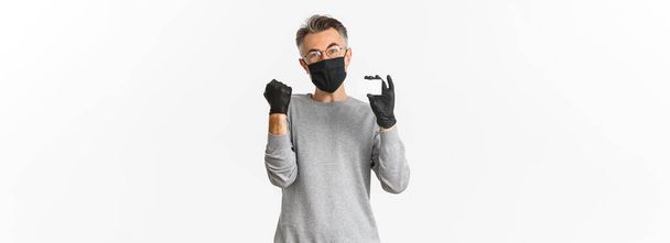 Concept of covid-19, social distancing and lifestyle. Image of attractive middle-aged man in medical mask, gloves and glasses, showing credit card and rejoicing, standing over white background. - Photo, Image