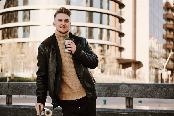 Portret białego młodzieńca z deskorolką pijącego kawę na ulicy - Zdjęcie, obraz