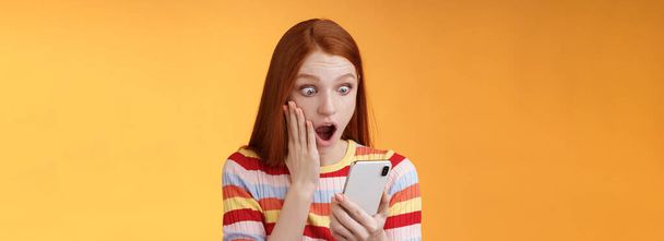 Omg what heck. Portrait shocked concerned young redhead sensitive impressed redhead woman stare smartphone display touch cheek drop jaw stunned surprised standing orange background hold phone. - Photo, Image