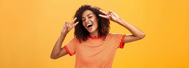 Nobody gonna spoil my perfect mood. Portrait of happy optimistic attractive dark-skinned female model with curly hairstyle tilting head smiling carefree showing peace or victory signs near face. Copy - Photo, Image