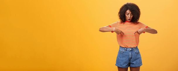 Retrato de preocupado questionado e surpreendido afro-americano feminino com penteado encaracolado olhando e apontando para o estômago ou barriga sentindo desconforto e problemas com a saúde sobre fundo laranja - Foto, Imagem