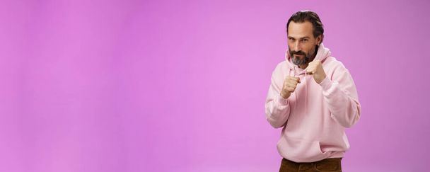 pyskaty pewny siebie dobrze wyglądający dojrzały kaukaski brodaty mężczyzna w różowej bluzie uczy syna jak stać z tyłu walczyć bronić się podnosić pięści gotowy boks uśmiechnięty śmiały, pozostać dobry kształt, fioletowy tło. - Zdjęcie, obraz