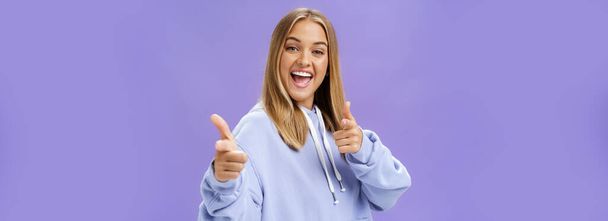 Enthusiastic and charismatic woman feeling joyful meeting friends checking out cool copy space pointing with finger guns at camera smiling broadly posing in stylish hoodie over purple wall. Copy space - Photo, Image