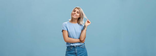 Студийный снимок расслабленной и спокойной популярной привлекательной женщины с светлыми волосами и татуировками, играющей с наклонной головой и улыбающейся, как будто слушающий бойфренд или флиртующий стоящий забавно над синей стеной - Фото, изображение