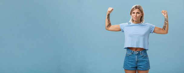 Стиль жизни. Сильные и сильные симпатичные спортсменки с татуировками в футболках и шортах, поднимающие руки и показывающие большие мускулы и бицепсы, гордо улыбающиеся, хвастающиеся физической силой над синим - Фото, изображение