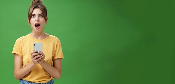 Portret zszokowanej niemową i pod wrażeniem pięknej białej dziewczyny z czesanymi włosami w żółtym t-shircie trzymającej smartfona, opadająca szczęka z podniecenia reagująca na chłodną aplikację na zielonym tle. - Zdjęcie, obraz