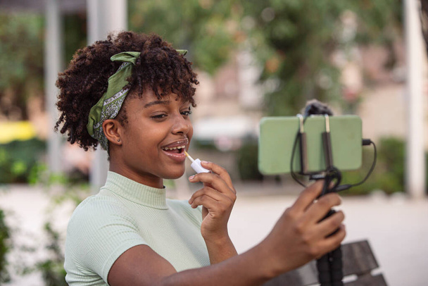 Sorrindo afro-latina estudante segurando smartphone, aprendizagem remota, chamada de vídeo smartphone sentado no banco ao ar livre - Foto, Imagem