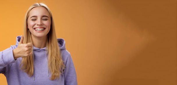 Amused joyful optimistic young excited blond european female model in hoodie smiling broadly close eyes having fun show approval gesture thumbs-up like awesome trip vacation idea, orange background. - Photo, Image
